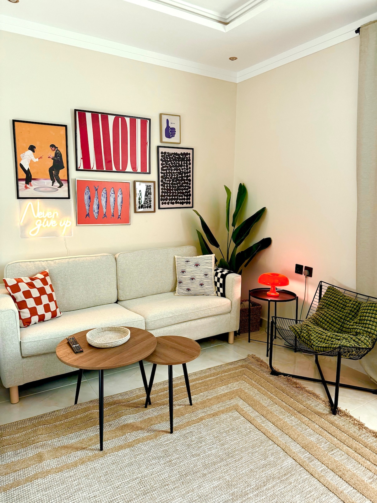 A cozy living area features a light-colored sofa adorned with colorful cushions. A set of round coffee tables is positioned in front, alongside a modern black wire chair. The wall displays an eclectic mix of art pieces and a neon sign, while a green plant adds a touch of nature.