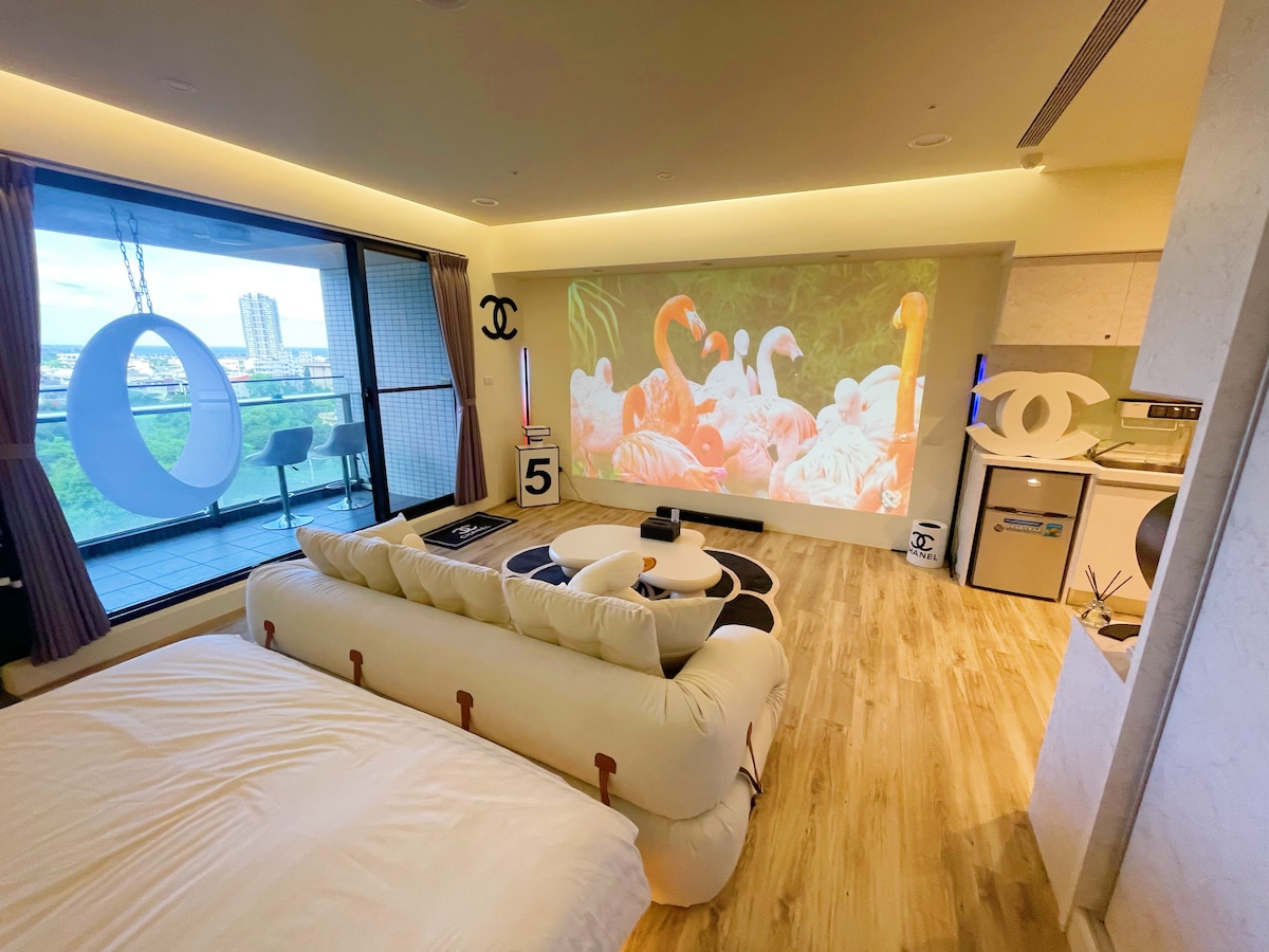 A spacious room features a large bed with a cozy white comforter. A projection of flamingos adorns one wall, enhancing the ambiance. A balcony with glass sliding doors opens to views of the city, while soft lighting creates a relaxing atmosphere.