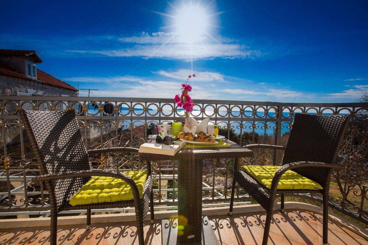 A balcony is presented with two woven chairs featuring bright green cushions positioned around a small table. A floral centerpiece adds a touch of color. The sun shines overhead, illuminating a view of the water and a clear blue sky.