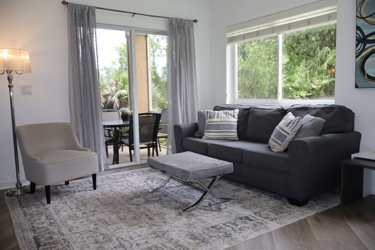 A cozy seating area features a gray sofa adorned with decorative pillows, situated on a patterned rug. A stylish armchair complements the space. Natural light enters through a large window, revealing a view of the outdoor seating area surrounded by greenery.