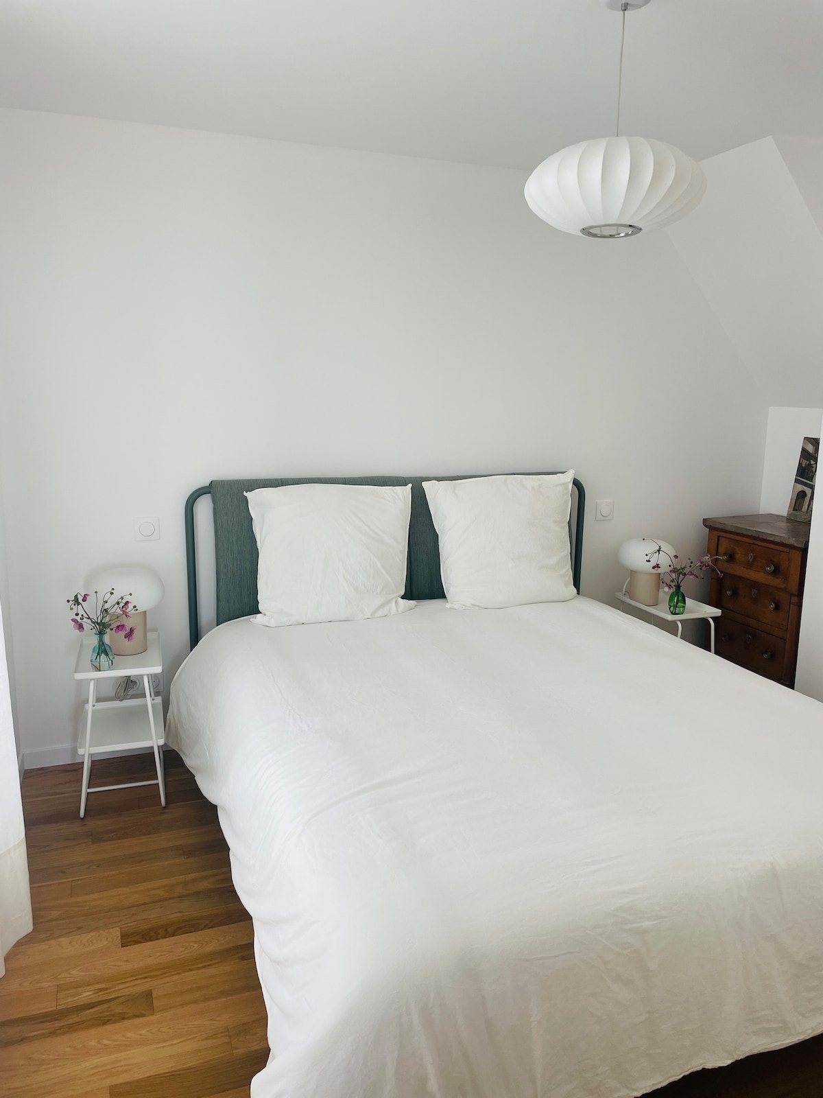 A serene bedroom features a soft white bedding on a spacious bed with two white pillows. On either side of the bed, small nightstands hold decorative lamps. A vintage wooden dresser adds character to the room, which is illuminated by natural light from the window.