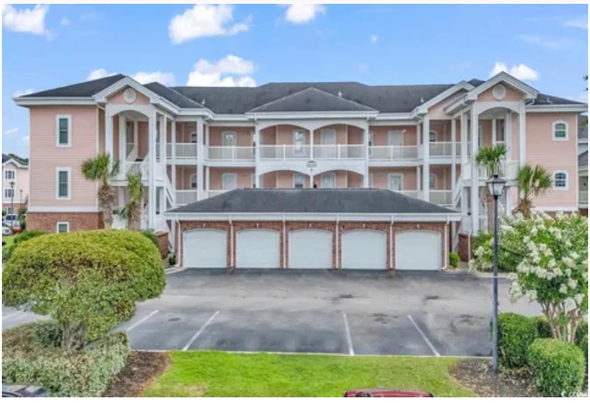 A two-story condo building features a soft pink exterior with white railings and balconies. Manicured shrubs and small trees frame the parking area in front. Multiple garage doors are visible beneath the elevated structure, providing a spacious feel to the surrounding area.