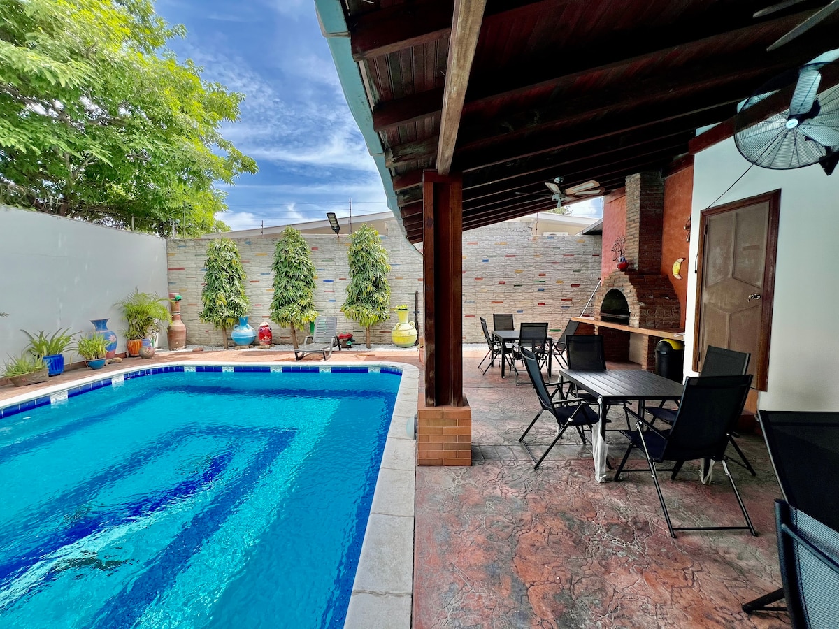 A swimming pool is surrounded by a spacious patio area featuring several tables and chairs. Lush greenery is visible along the walls, while a barbecue area is located to the right. A ceiling fan provides cooling air above the seating area.