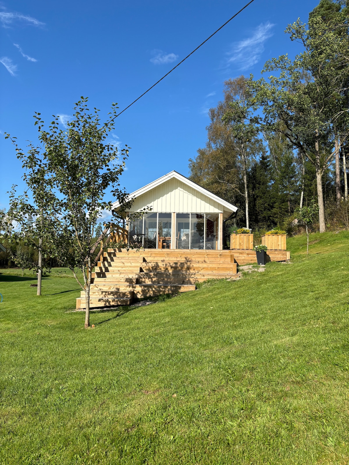 A charming house is set on a slight hill, surrounded by lush green grass and trees. A wooden deck extends from the glassed-in porch, providing outdoor seating. The clear blue sky enhances the serene atmosphere of this inviting retreat.