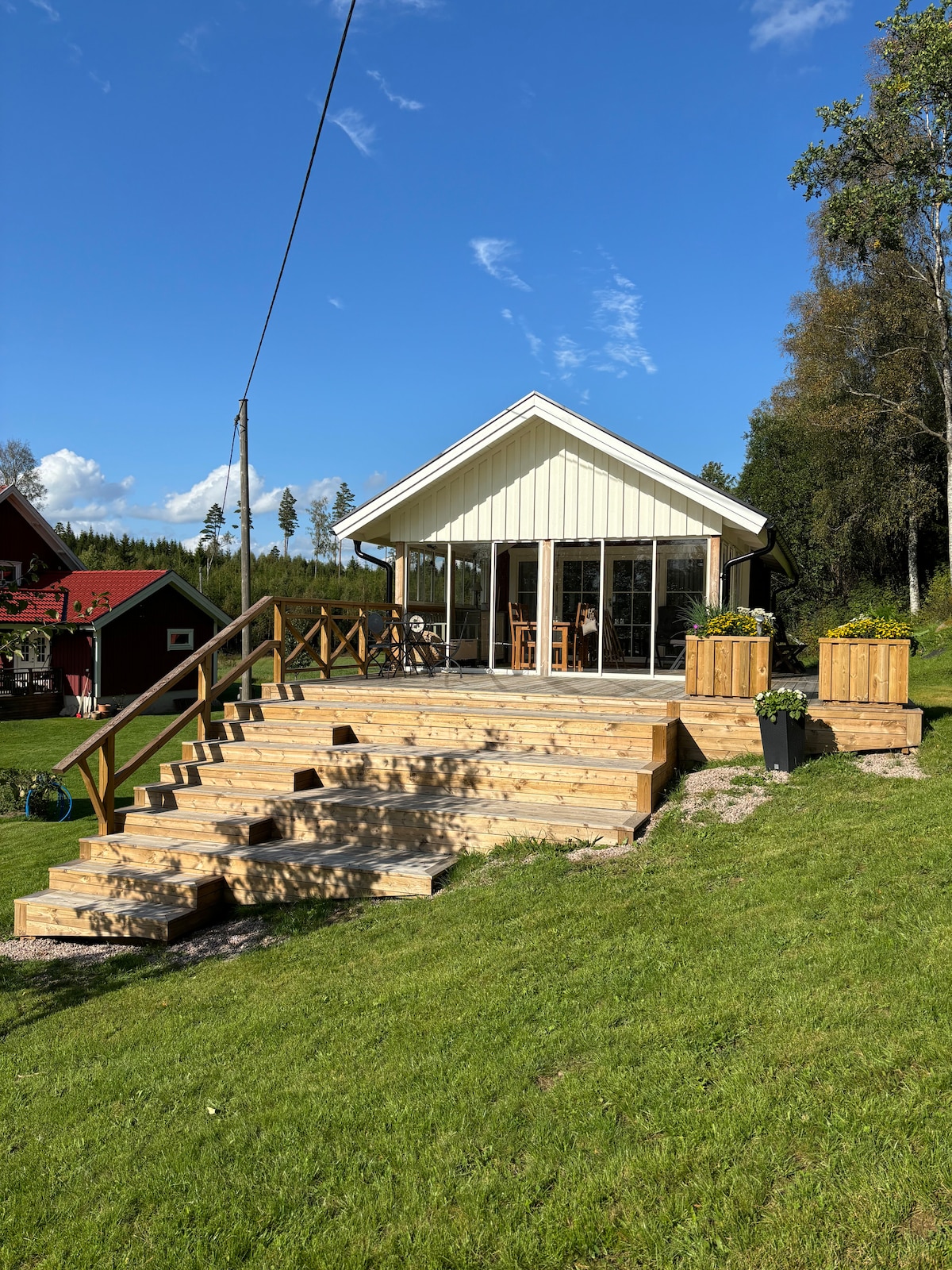 The exterior of a newly renovated cabin showcases a sunny facade with large glass doors that open to an expansive deck. Steps lead to a grassy area, surrounded by planters with greenery, providing an inviting entrance to the peaceful retreat.