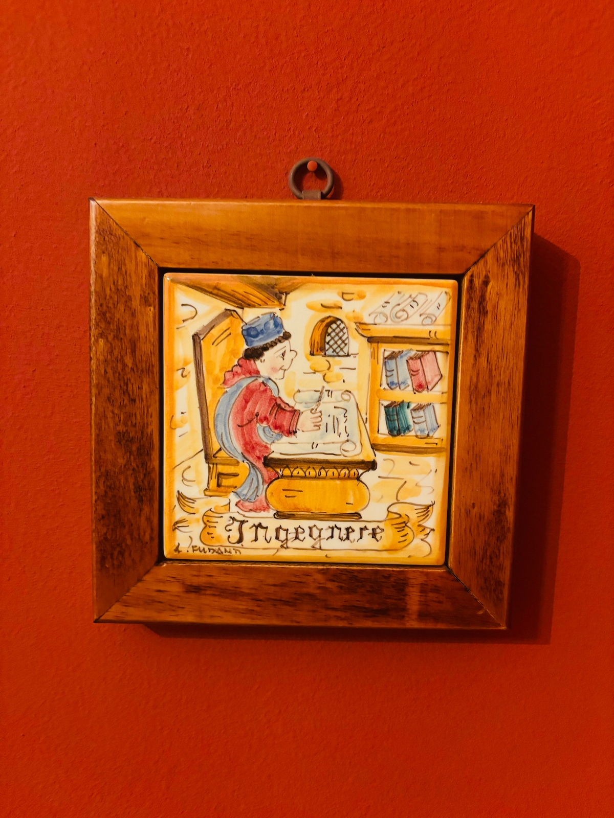 A framed piece of ceramic art is displayed against a red wall. The artwork depicts a figure in period attire, seated at a wooden desk surrounded by books. The frame is made of wood, providing a warm contrast to the vivid background.