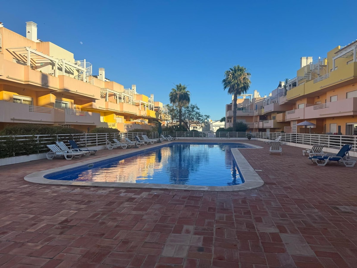 A tranquil outdoor pool area is framed by surrounding residences. Lounge chairs are placed along the poolside, and palm trees provide a touch of greenery. The clear blue sky reflects off the water, creating a serene atmosphere in the spacious communal area.