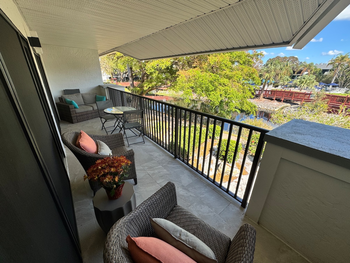 A private covered lanai patio is visible, featuring comfortable seating arranged around a small table. Lush greenery and a lagoon can be seen in the background, enhancing the serene outdoor atmosphere. The area is well-lit with natural light.