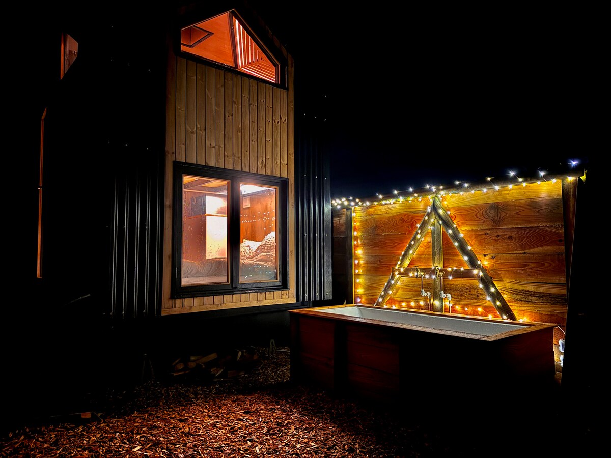 A cozy outdoor bath area is featured alongside a contemporary structure, illuminated by string lights enhancing the wooden paneling. A large window reveals a softly lit interior with a spacious bed, inviting a sense of comfort and relaxation.