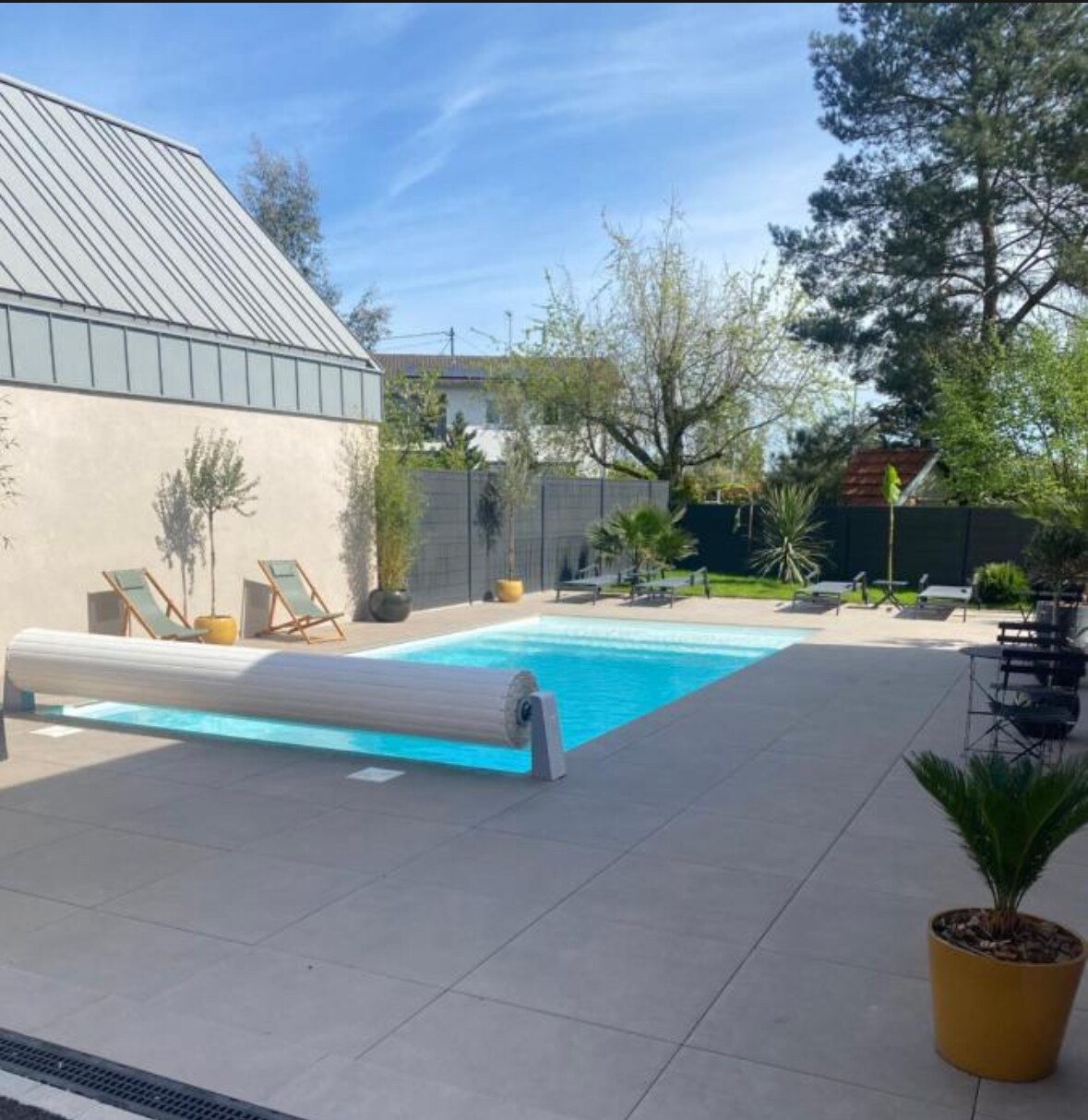 An outdoor area features a heated swimming pool surrounded by modern stone tiles. Sun loungers are placed beside the pool, with potted plants adding greenery. Fencing provides privacy, while trees and shrubs create a serene atmosphere.