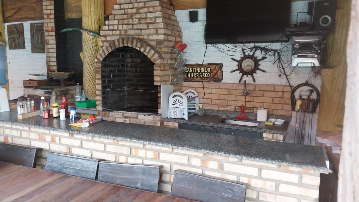 An outdoor kitchen space features a traditional brick barbecue grill, surrounded by a granite countertop. Various condiments and cooking utensils are neatly arranged on the counter. A wall-mounted television and decorative wall art complement the setup, creating an inviting environment for meal preparation.