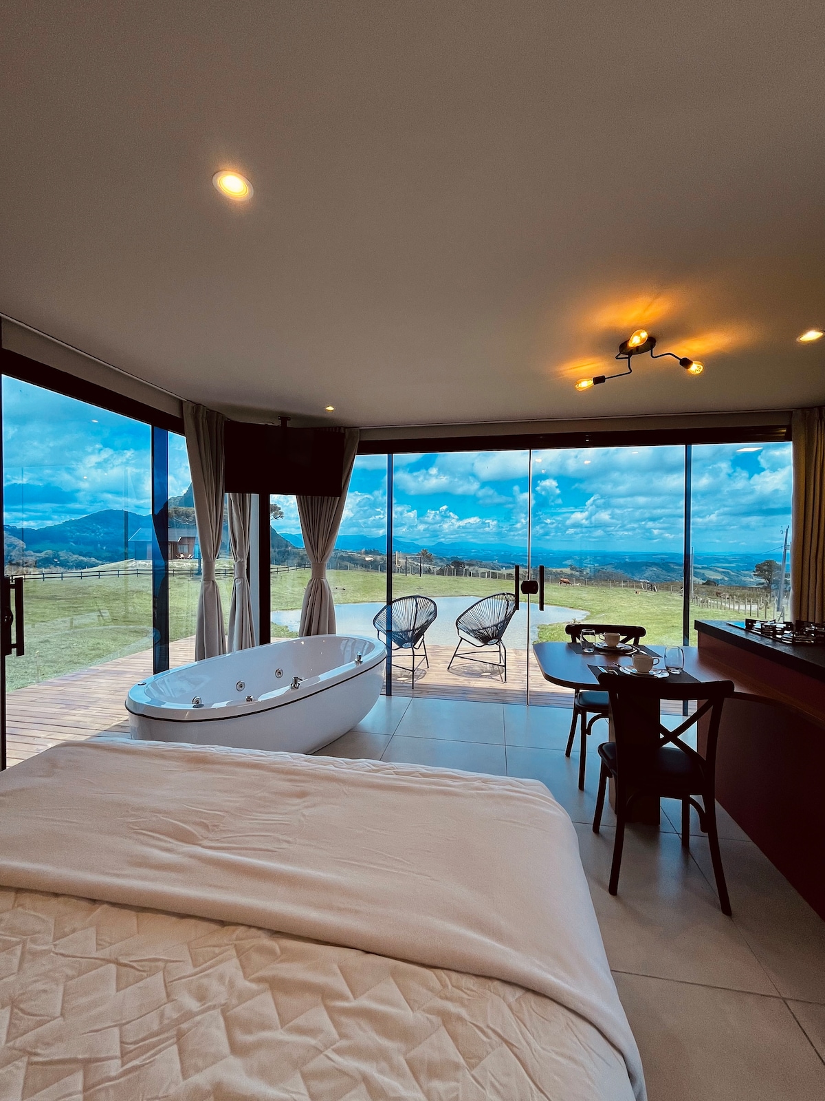A spacious interior includes a luxurious Jacuzzi tub near floor-to-ceiling windows, revealing expansive mountain views. A comfortable queen bed is positioned thoughtfully, with a compact dining table and chairs nearby. Natural light fills the room, enhancing the serene atmosphere.