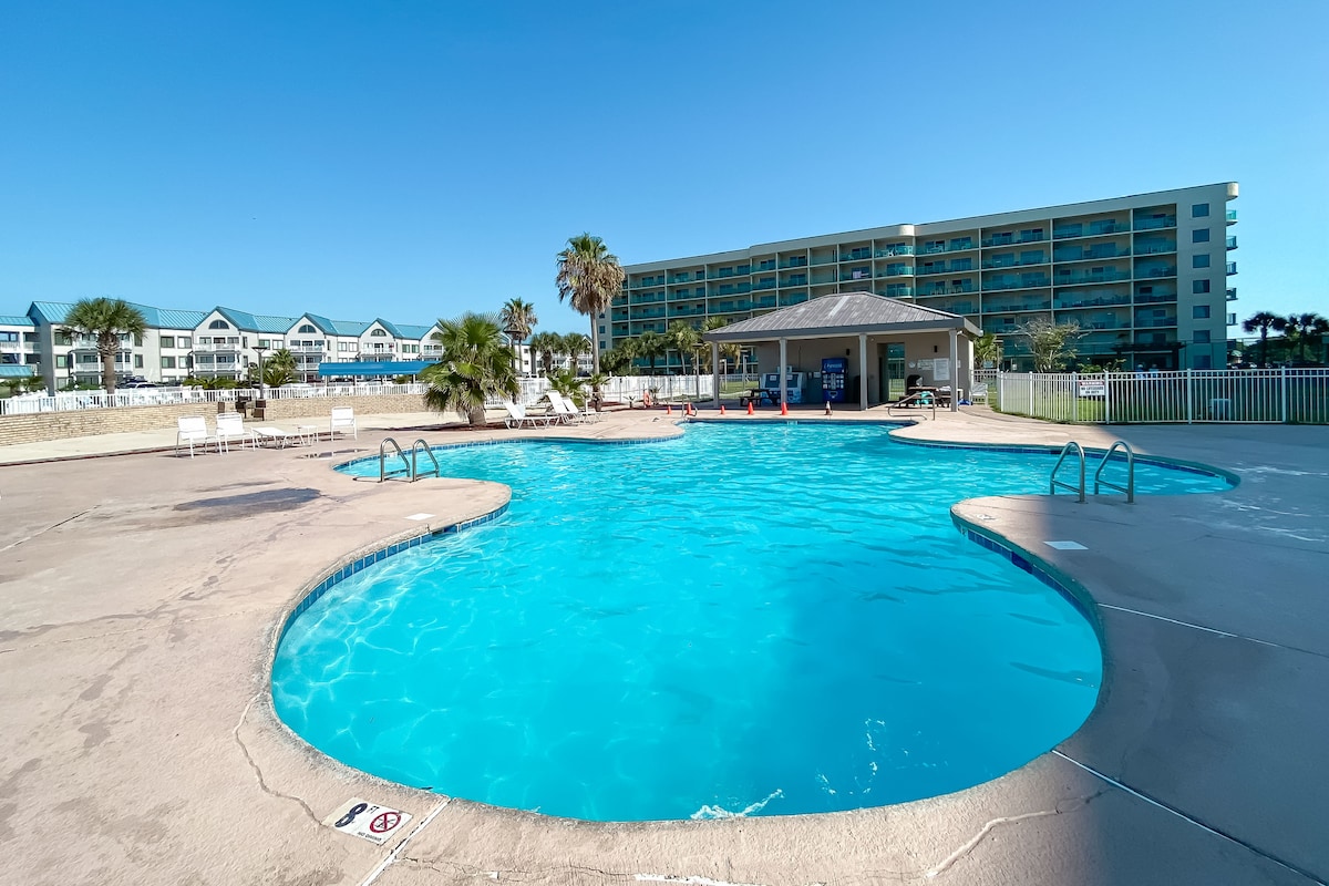An outdoor pool is shown, featuring a spacious curved design with clear blue water. Surrounding palm trees and lounge chairs create a relaxed atmosphere. In the background, the condo building can be seen, providing a bright backdrop against the clear sky.