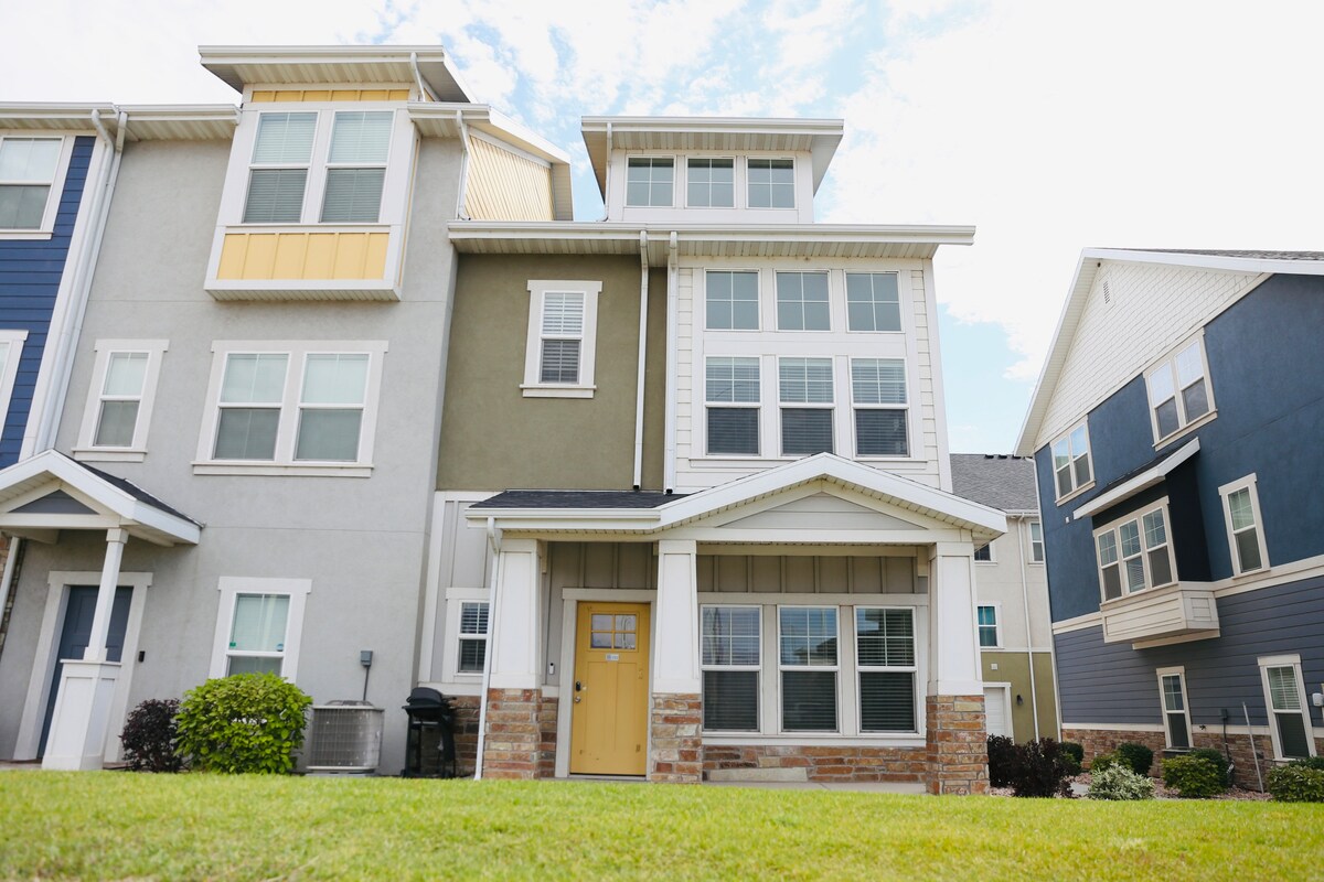 The exterior showcases a multi-level townhome with a prominent entrance featuring a yellow door. Large windows provide ample natural light. The building is set against a well-maintained lawn, with nearby greenery adding to the appealing facade.