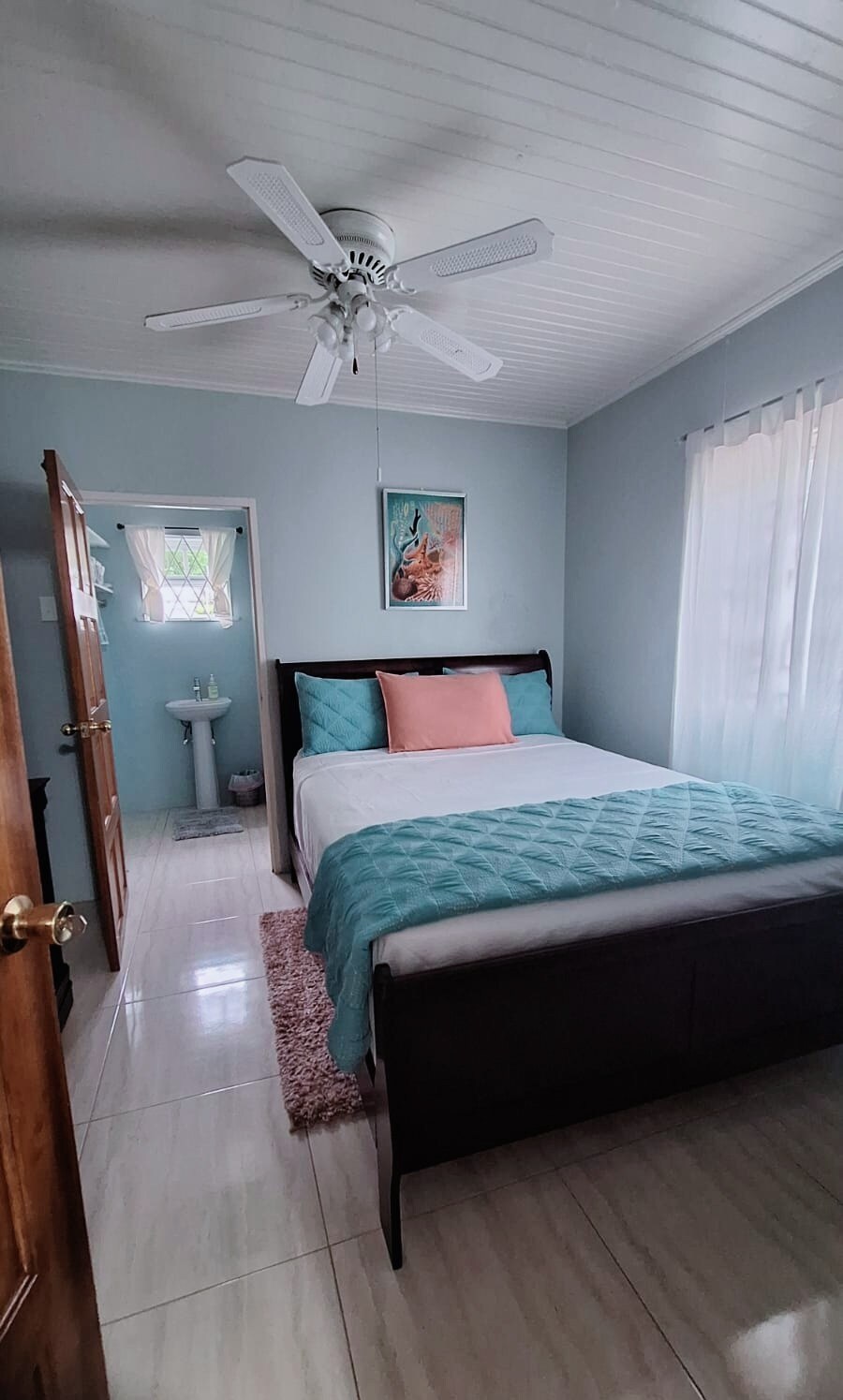 A bright bedroom is furnished with a dark wood bed dressed in a quilted blue coverlet and a decorative pink pillow. Natural light enters through two windows, complementing the soft blue walls. A small washbasin is visible in the corner, enhancing functionality.