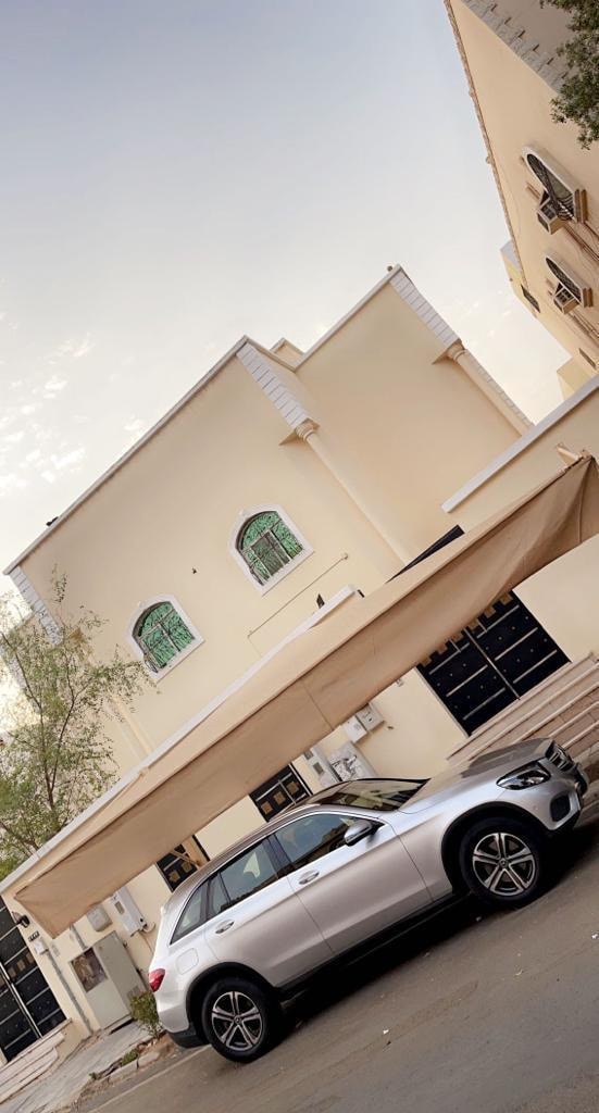 A spacious cream-colored building is shown, featuring multiple windows that allow natural light. A sleek silver car is parked under a protective canopy in front, while the surrounding area includes sidewalks and greenery, enhancing the inviting layout.