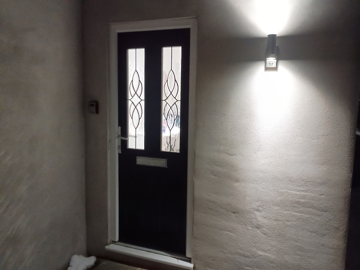 A private entrance is visible, featuring a solid dark door with decorative glass panels. A soft outdoor light illuminates the entrance area, which is framed by a smooth, light-colored wall. Snow can be seen on the ground, adding a seasonal touch.