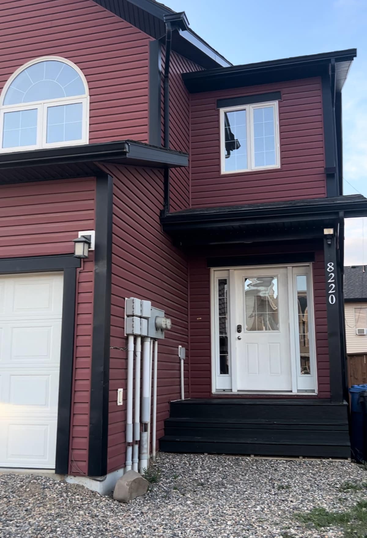 The exterior of the home features rich burgundy siding complemented by black accents. A covered porch with a white door leads into the house, accompanied by large windows that allow natural light to enter. A driveway and garage area are visible beside the entrance.