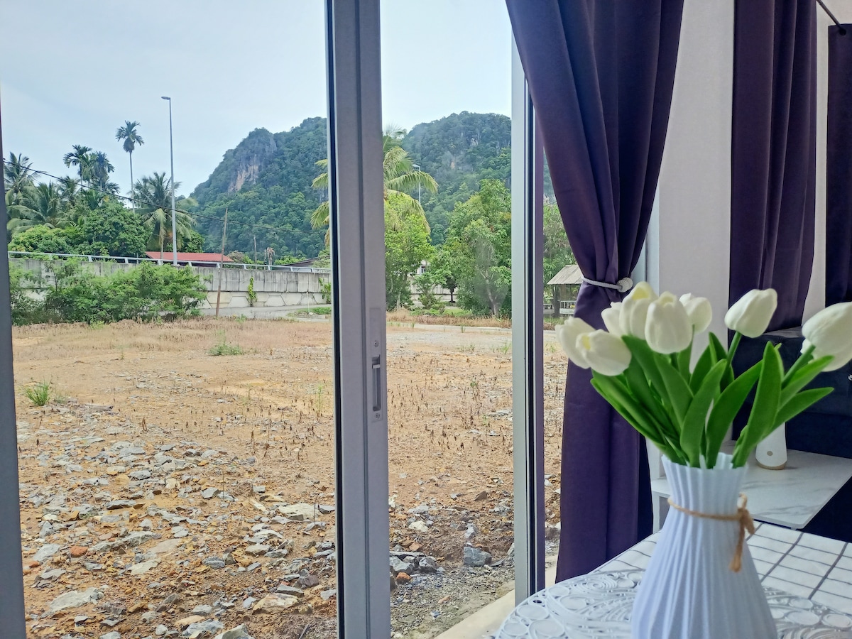 A view of the surrounding landscape is captured through large glass windows, revealing a stretch of land featuring uneven terrain and scattered stones. Lush greenery and a mountainous backdrop can be seen in the distance, while a vase of white tulips decorates the nearby table.