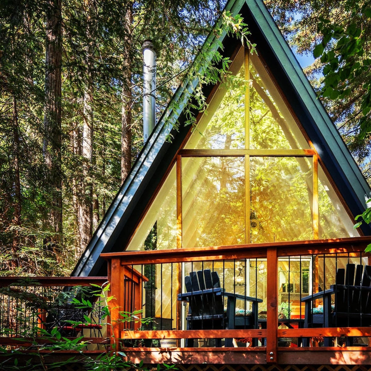 An A-frame cabin is surrounded by tall trees, featuring a large glass front that invites natural light. A wooden deck with two adirondack chairs offers a cozy outdoor seating area, inviting relaxation in a serene setting.