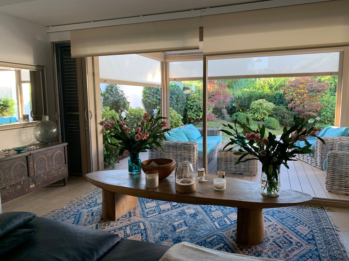 A spacious living area is highlighted by large glass doors that open to a serene garden. Elegant floral arrangements sit on a wooden coffee table, while comfortable wicker seating is positioned nearby. Soft natural light filters through sheer window coverings, enhancing the tranquil setting.