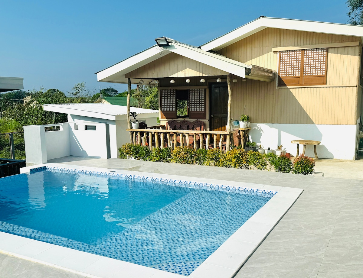 A modern bahay kubo is featured adjacent to a clear blue pool, surrounded by landscaped greenery. A spacious patio with wooden accents is visible, offering comfortable seating areas and large windows that open to the vibrant outdoor scenery.