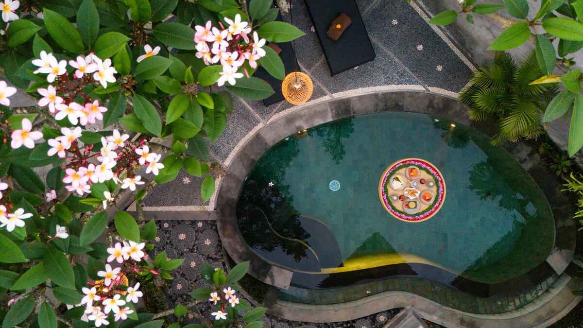 An aerial view captures a tranquil swimming pool surrounded by lush greenery and blooming flower trees. The pool features a circular arrangement of colorful floats, inviting relaxation. The stone pathway leading to the pool is visible, enhancing the serene environment.