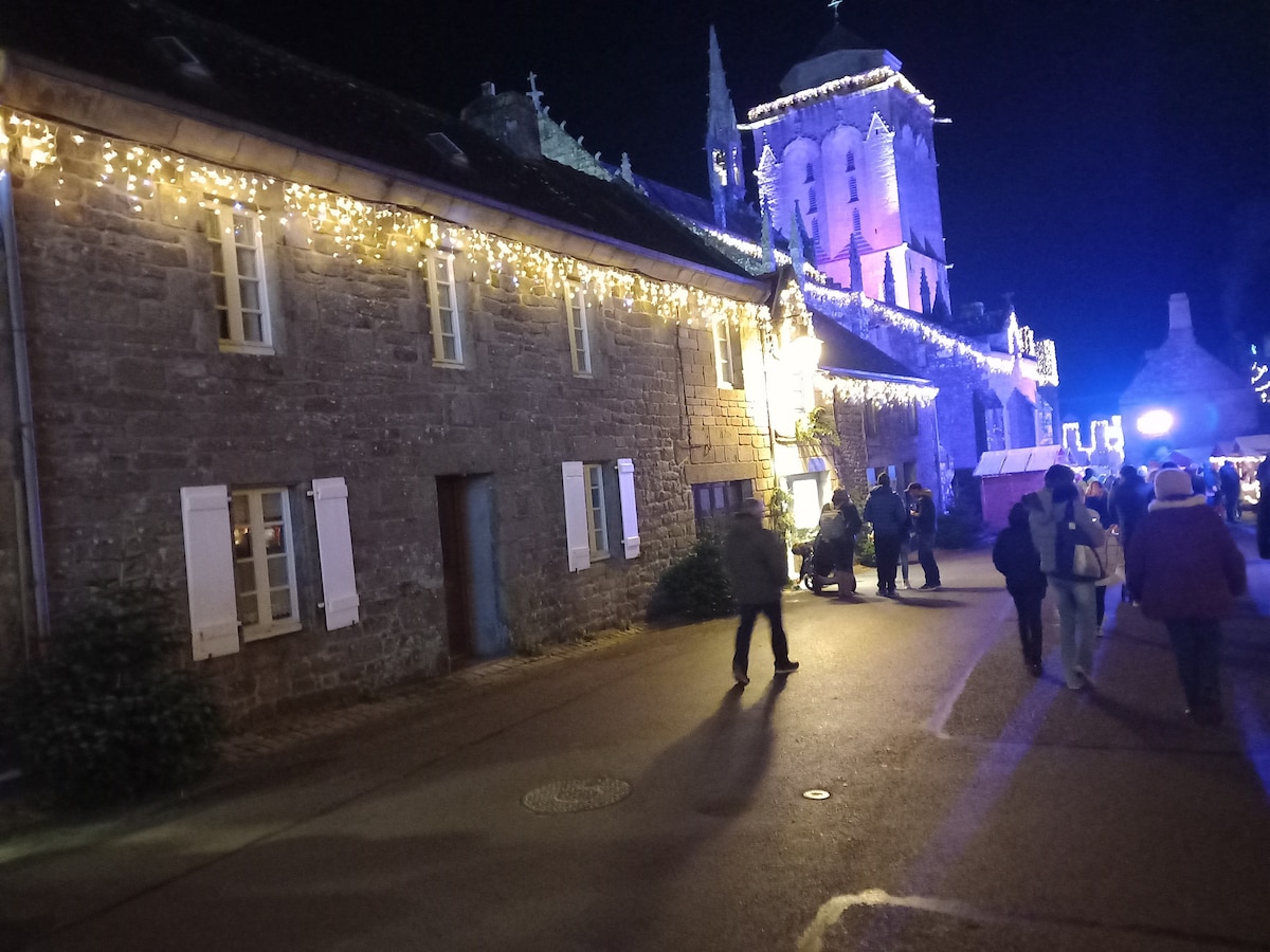 A charming building is illuminated by soft white lights, highlighting its stone façade and traditional shutters. The scene is lively, with people gathered on the street, adding to the vibrant community atmosphere. Warm light spills from the windows, hinting at the inviting interiors.