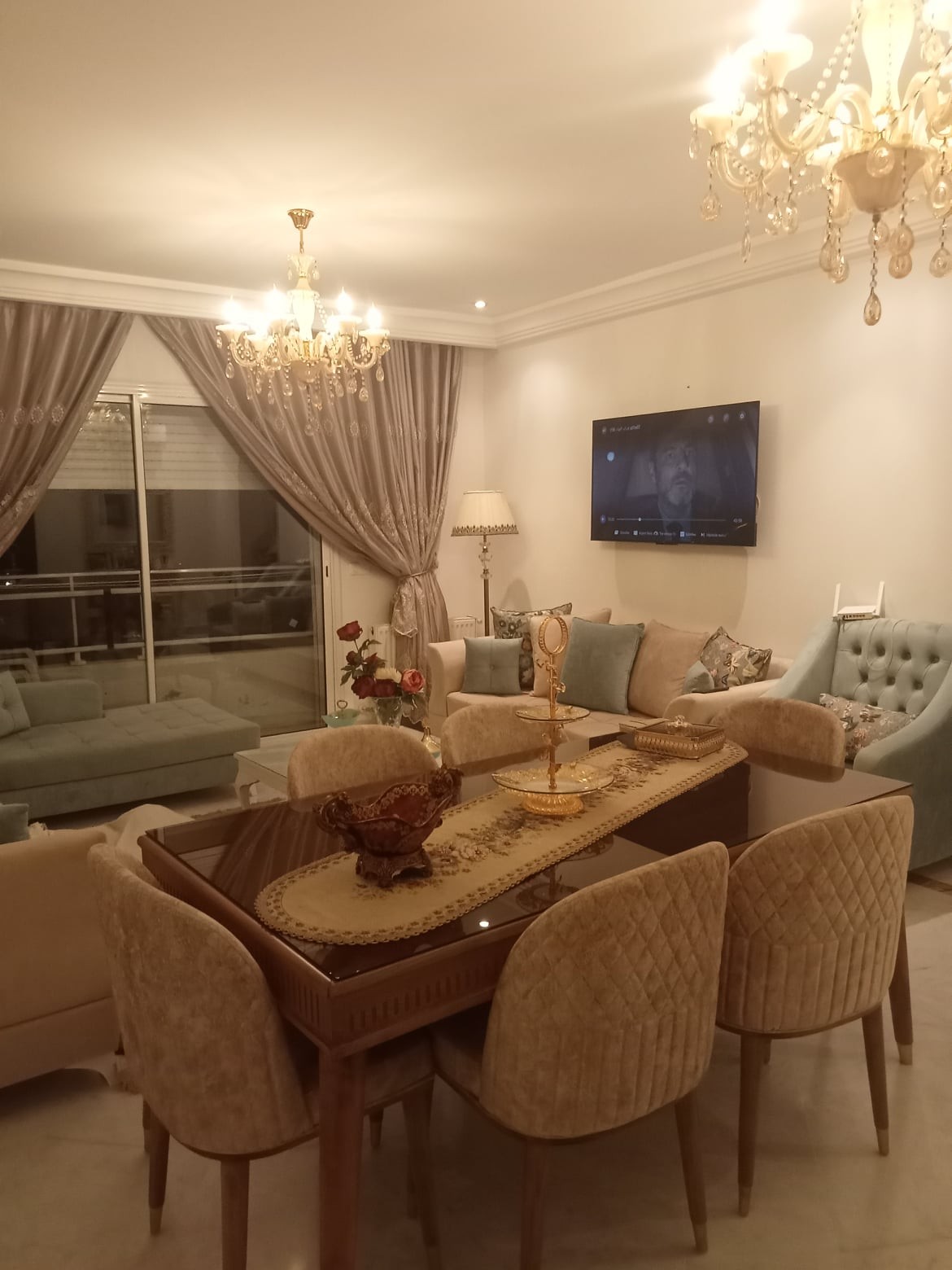 An elegant dining area is displayed, featuring a polished wooden table surrounded by upholstered chairs. Soft lighting from a chandelier casts a warm glow, complemented by neutral-toned curtains and decorative accents. A comfortable seating area is visible in the background, enhancing the inviting ambiance.