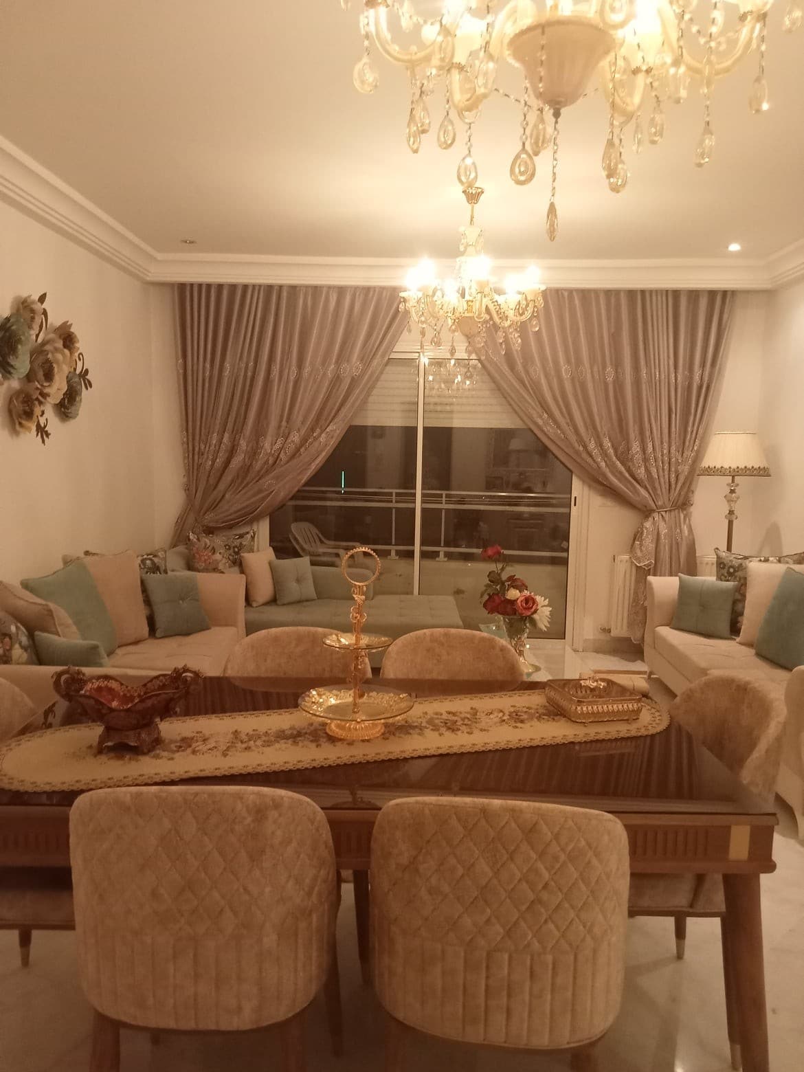 A stylish living and dining area is presented, featuring a large polished dining table surrounded by upholstered chairs. Soft lighting is provided by a chandelier, and elegant drapes frame the windows. Decorative pieces are arranged on the table, enhancing the sophisticated ambiance.