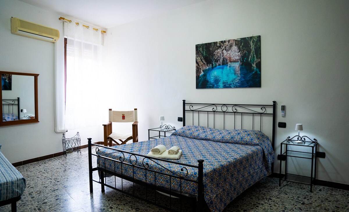 A spacious bedroom features a large bed with a patterned blue coverlet. An image of a turquoise pool adorns the wall above the bed. Two nightstands and a mirror are situated beside the bed, while a wooden chair is positioned in the corner.