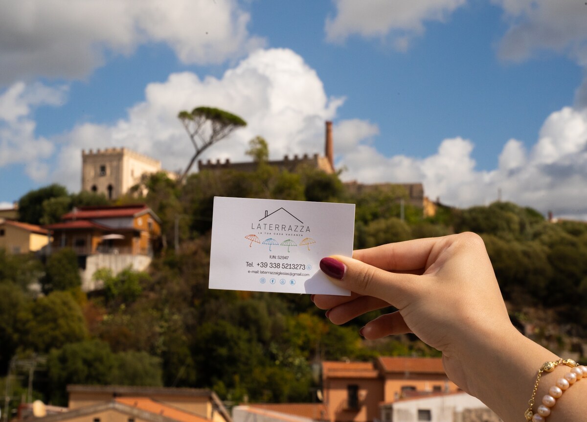 A hand holds a business card showcasing the name and contact information of the property, with a backdrop of a historic building and greenery under a partly cloudy sky. The scene captures the surrounding area, emphasizing its proximity to local attractions.