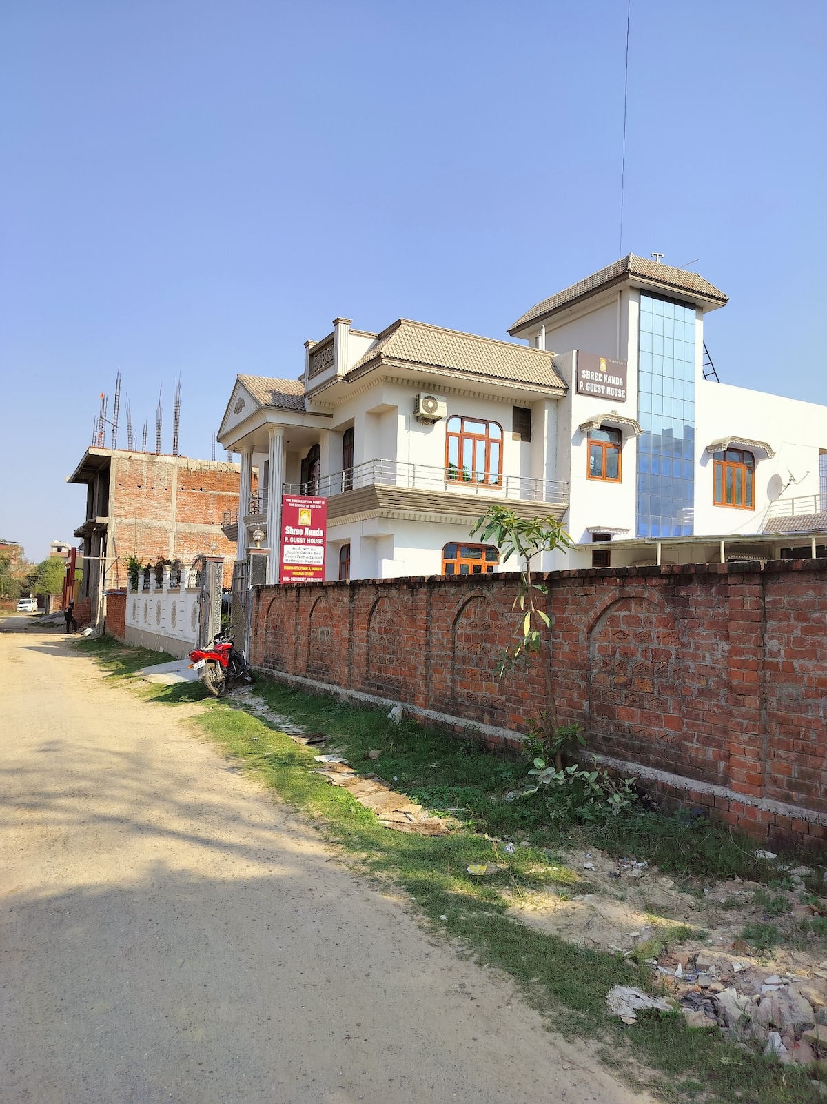 A large, modern building is set along a tree-lined street. The structure displays a combination of white and glass elements along with traditional detailing. A brick wall borders the property, with greenery visible along the path, contributing to the spacious outdoor setting.