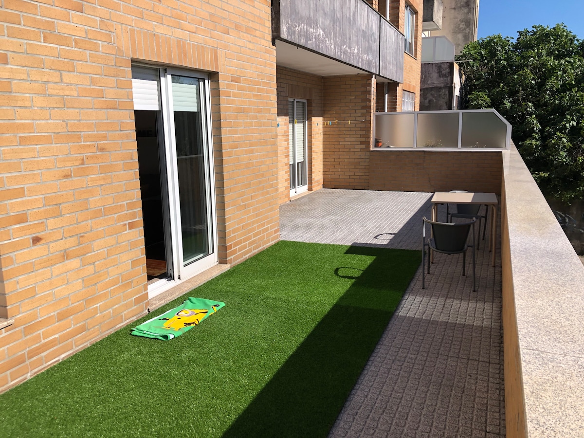 A private terrace is shown with artificial grass, offering a green space. A small table and chairs are positioned near the wall, while sliding glass doors lead into the interior of the home. A view of greenery is visible in the background.