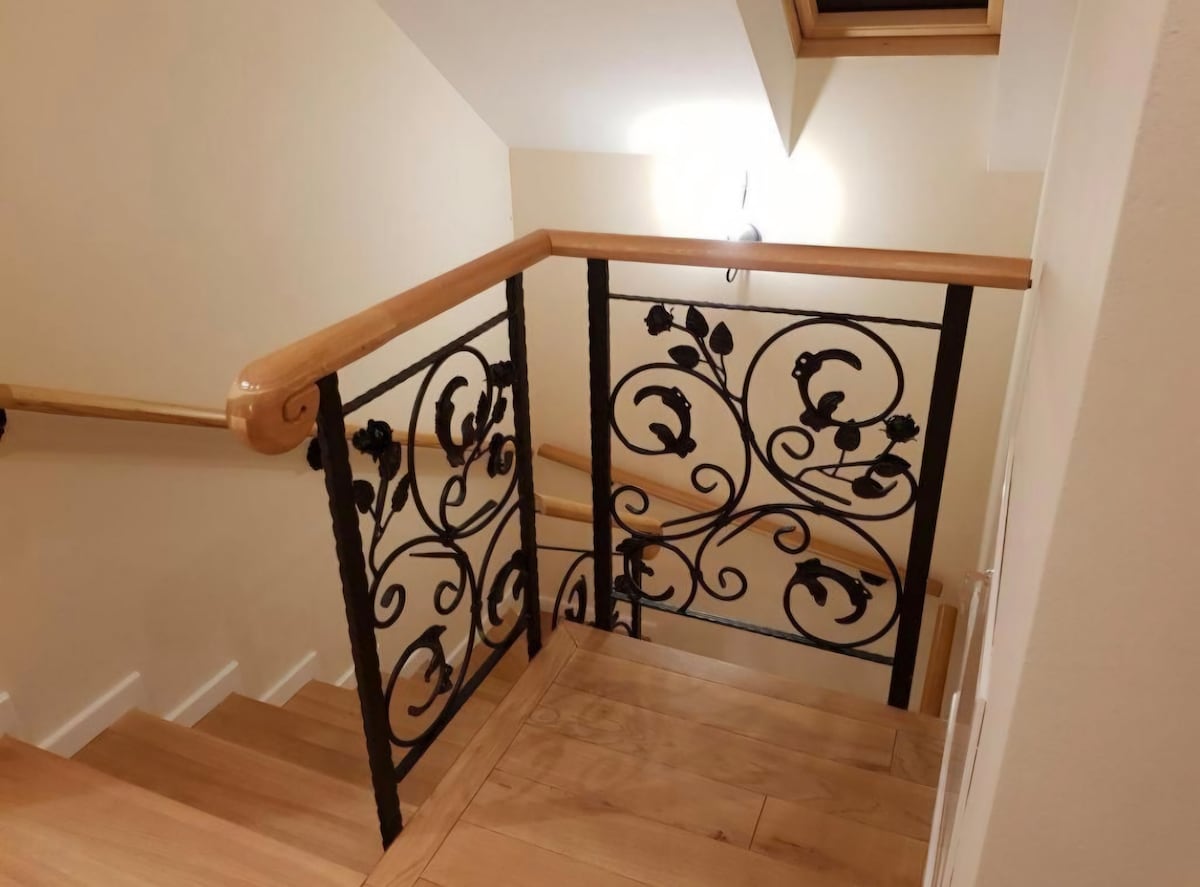 A well-crafted wooden staircase is seen leading downwards, framed by an ornate metal railing featuring floral designs. The staircase is illuminated by a soft light fixture on the wall, giving a warm appearance to the polished wooden steps.