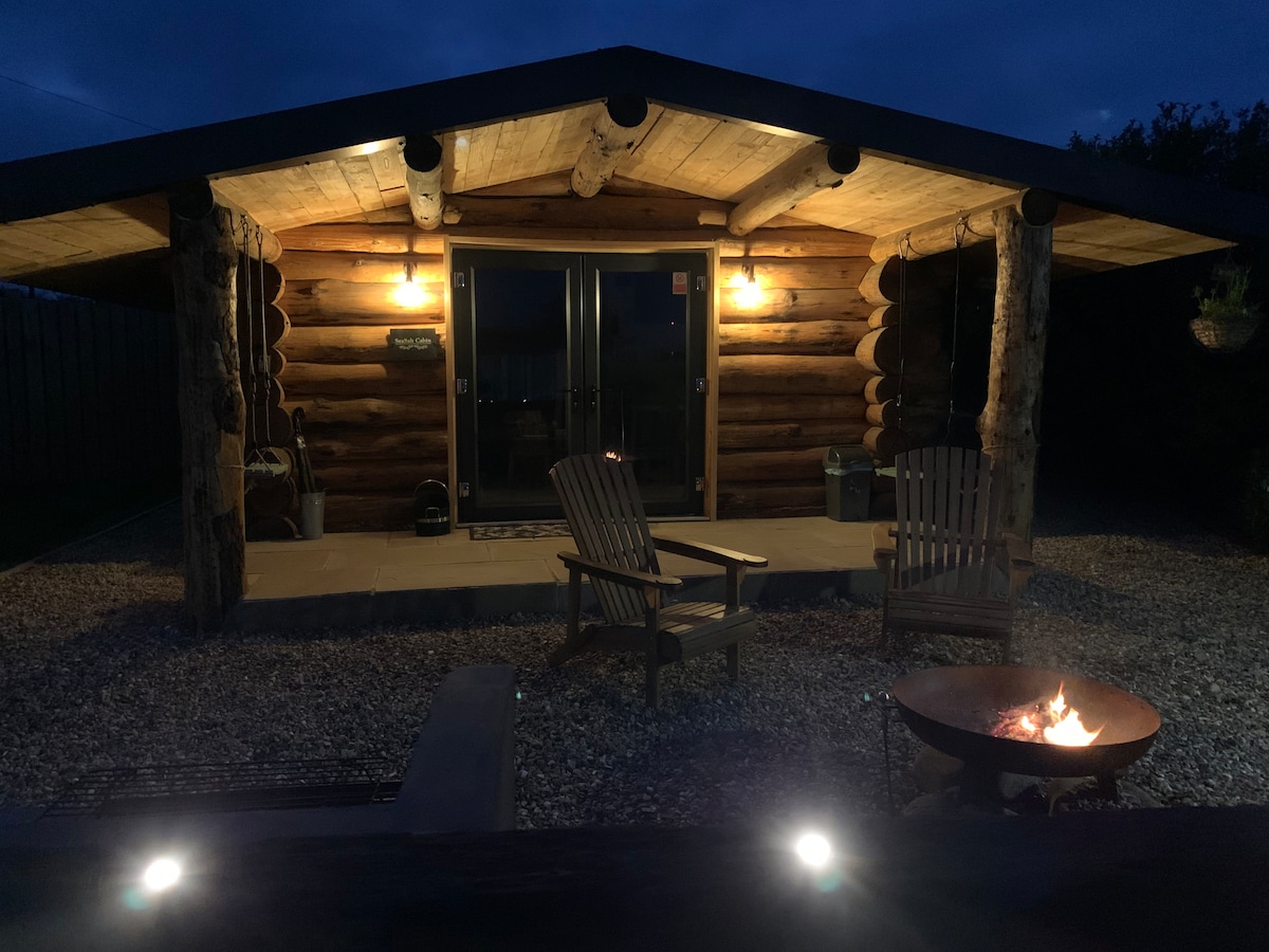 The log cabin exterior is softly illuminated at night, featuring wooden beams and stone flooring. Two wooden chairs are positioned near a fire pit, creating a cozy outdoor gathering space. Soft glowing lights accentuate the entrance, enhancing the welcoming ambiance.