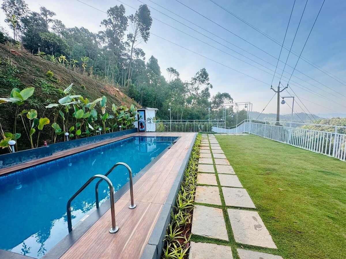 A serene swimming pool is surrounded by lush greenery, with stepping stones leading across well-maintained grass. The area showcases a view of distant hills under a clear sky, emphasizing the tranquil outdoor environment.