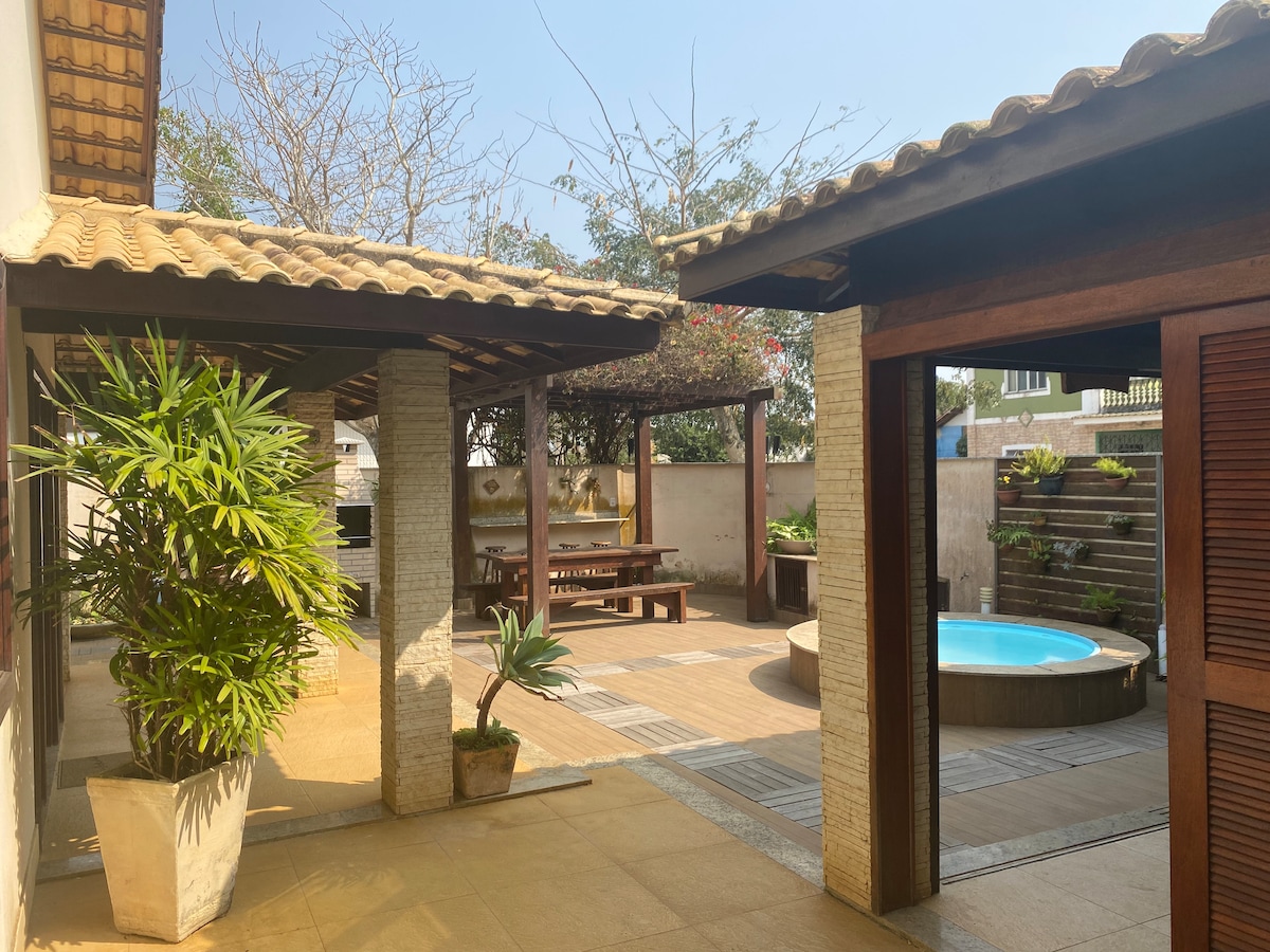 An outdoor space is framed by wooden structures, featuring a dining area with a large wooden table and benches. A palm plant is positioned in a pot, while a circular pool is visible in the background. The patio is illuminated by natural light, enhancing the area’s inviting ambiance.