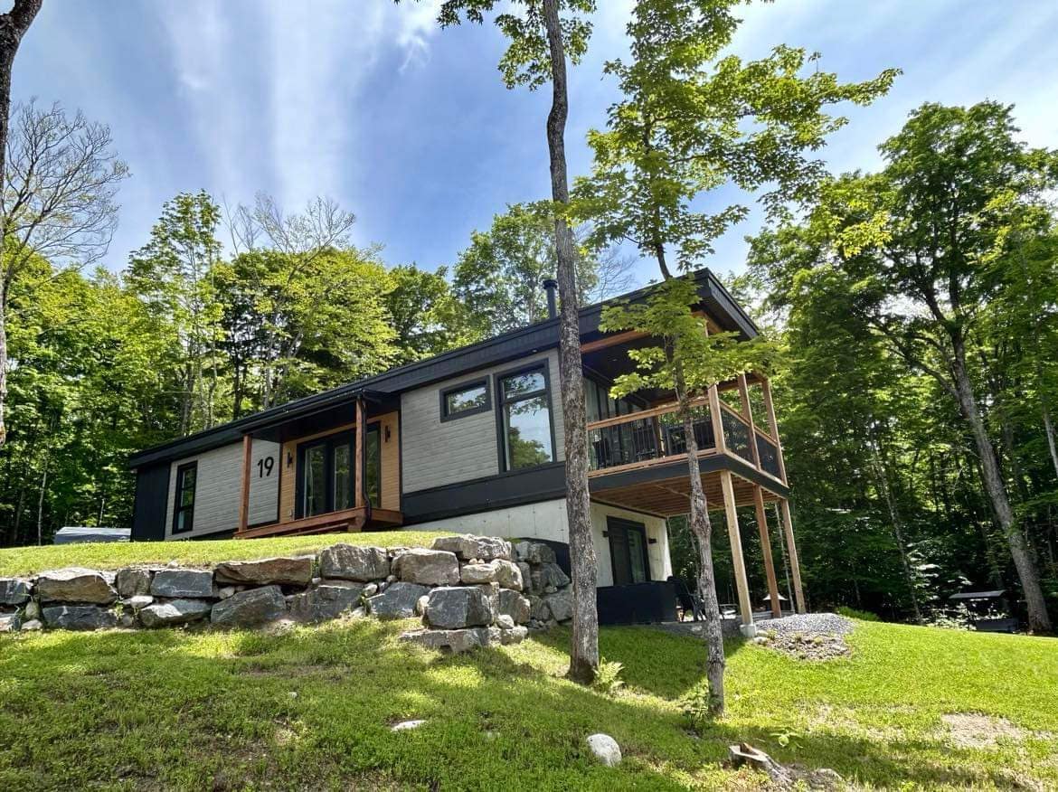 A modern chalet is set amidst lush greenery, showcasing a two-story design with a blend of wooden and stone elements. The entrance is accentuated by a stone pathway and a wooden deck. Sunlight filters through the trees, enhancing the natural landscape.