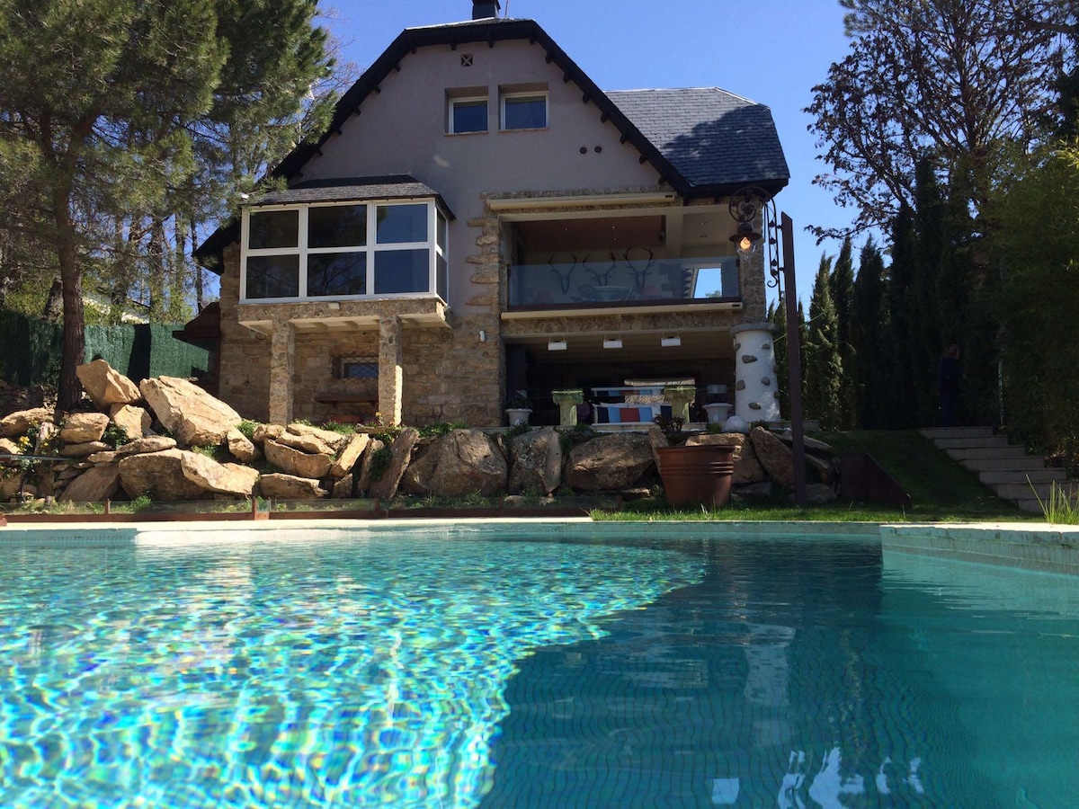 The exterior view of a two-story house is highlighted, featuring a combination of stone and modern elements. A glistening swimming pool occupies the foreground, reflecting clear blue skies, while a lush garden surrounds the property. The structure is framed by trees, adding to its natural setting.