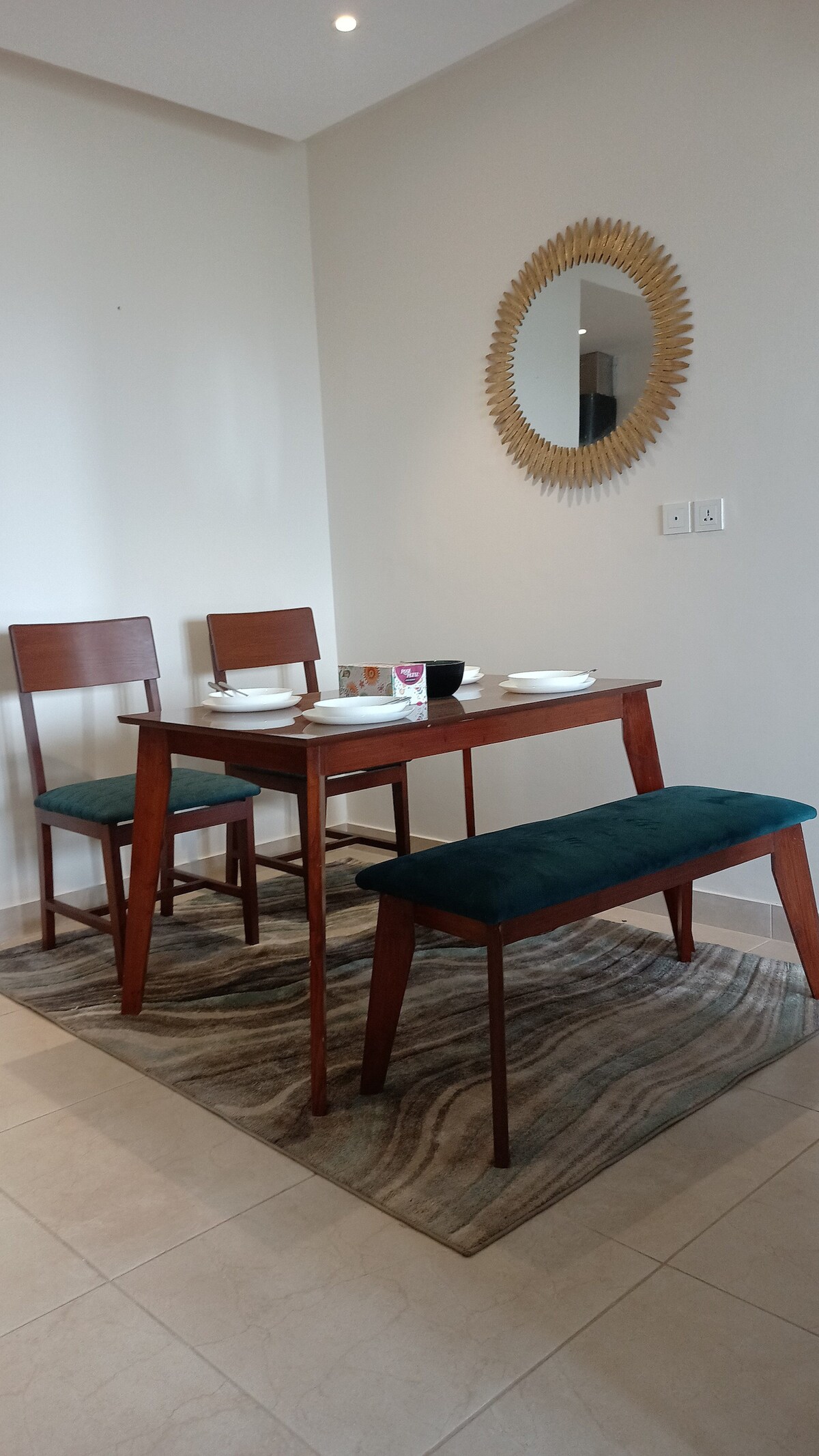 A dining area features a wooden table with matching chairs and a bench. Plates and cups are neatly arranged on the table. A round mirror is mounted on the wall, reflecting ambient light. The dining space is complemented by a textured area rug.