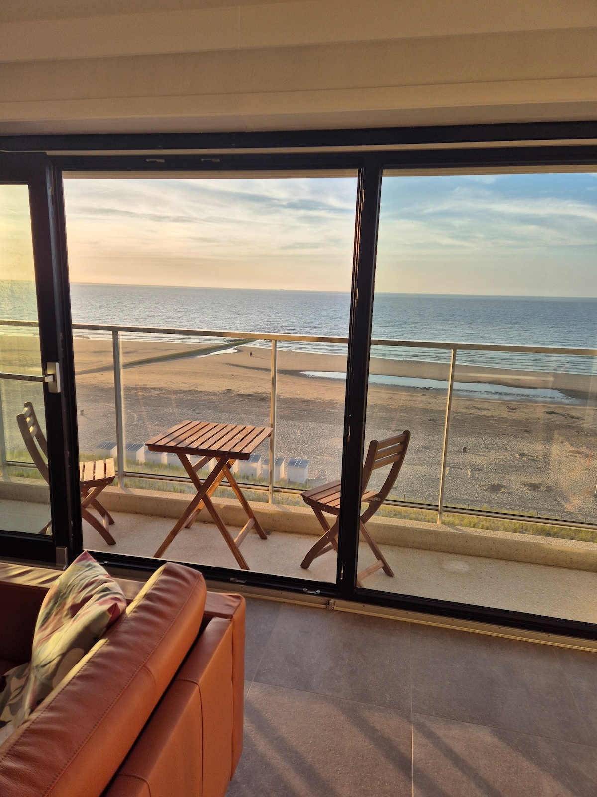 A large glass balcony door frames the serene view of the beach and ocean. A small wooden table is accompanied by two chairs on the balcony, capturing the atmosphere of morning light reflecting off the water.