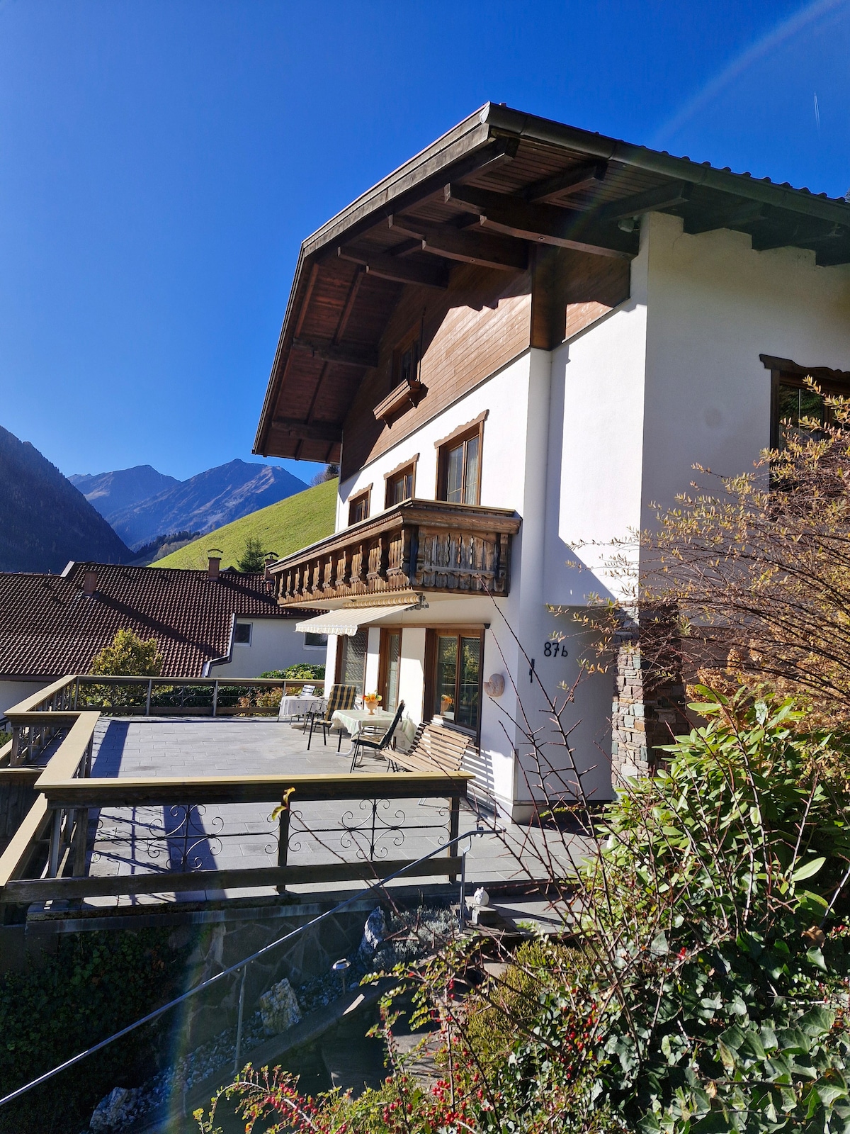A charming two-story house is set against a backdrop of mountains, featuring a large balcony with outdoor seating. The façade displays a combination of wood and white walls, and the surrounding greenery adds to the serene setting.
