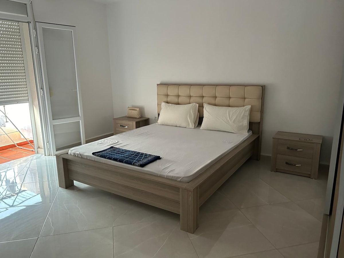 A spacious sleeping area features a light-colored upholstered bed with a neutral duvet. Flanking the bed are two wooden nightstands. Natural light enters through sliding glass doors that lead to a balcony, set against a wall of smooth white tiles.
