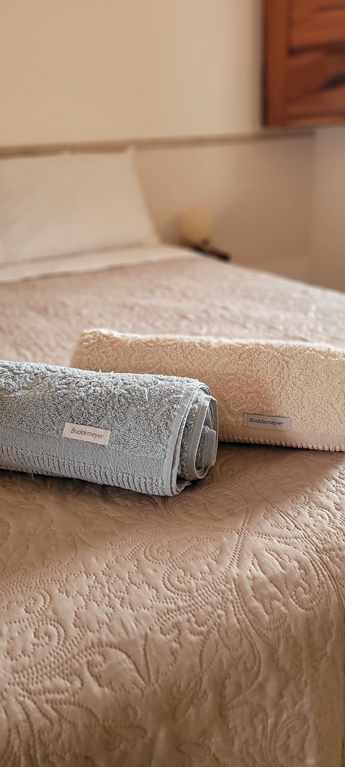 Two neatly rolled towels are placed on a textured bedspread, showcasing a soft blue towel alongside a cream-colored one. The spacious bed is made up with pristine linens, highlighting a tranquil ambiance in the room.