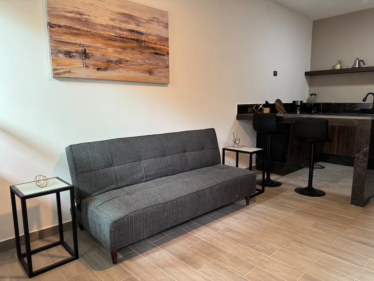 A gray sofa bed is positioned against a light wall, accompanied by two minimalist side tables. A landscape painting hangs above the sofa. In the background, a compact kitchen area features dark cabinetry and bar seating with two stools.