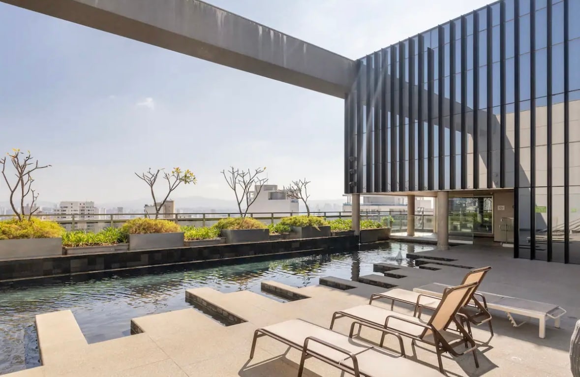 An outdoor terrace features a relaxing pool with clean lines, complemented by lounge chairs positioned nearby. Greenery adorns the perimeter, with sparse trees providing a touch of nature. The modern building facade stands prominently against the sky, reflecting a harmonious blend of architecture and landscape.