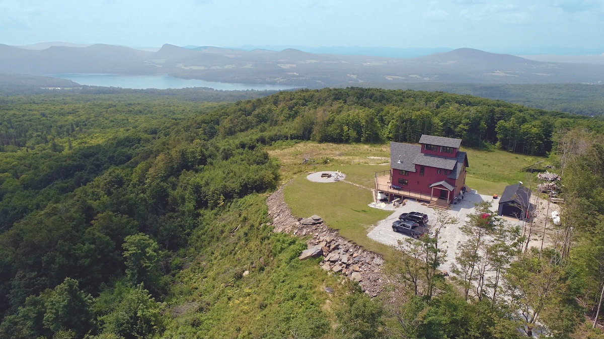 An aerial view depicts a multi-level red house situated on a hillside, surrounded by lush greenery. Scenic landscapes extend across the horizon, with visible water features and mountain ranges in the background. A circular patio is observed in the foreground, accompanied by a gravel area for parking.