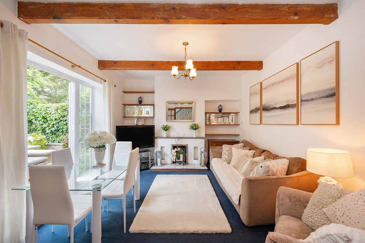 A bright living area features soft beige sofas, a glass dining table, and a cozy area rug. Natural light floods through large doors that lead to an outdoor space. Warm wooden beams and tasteful wall art add character to the space.