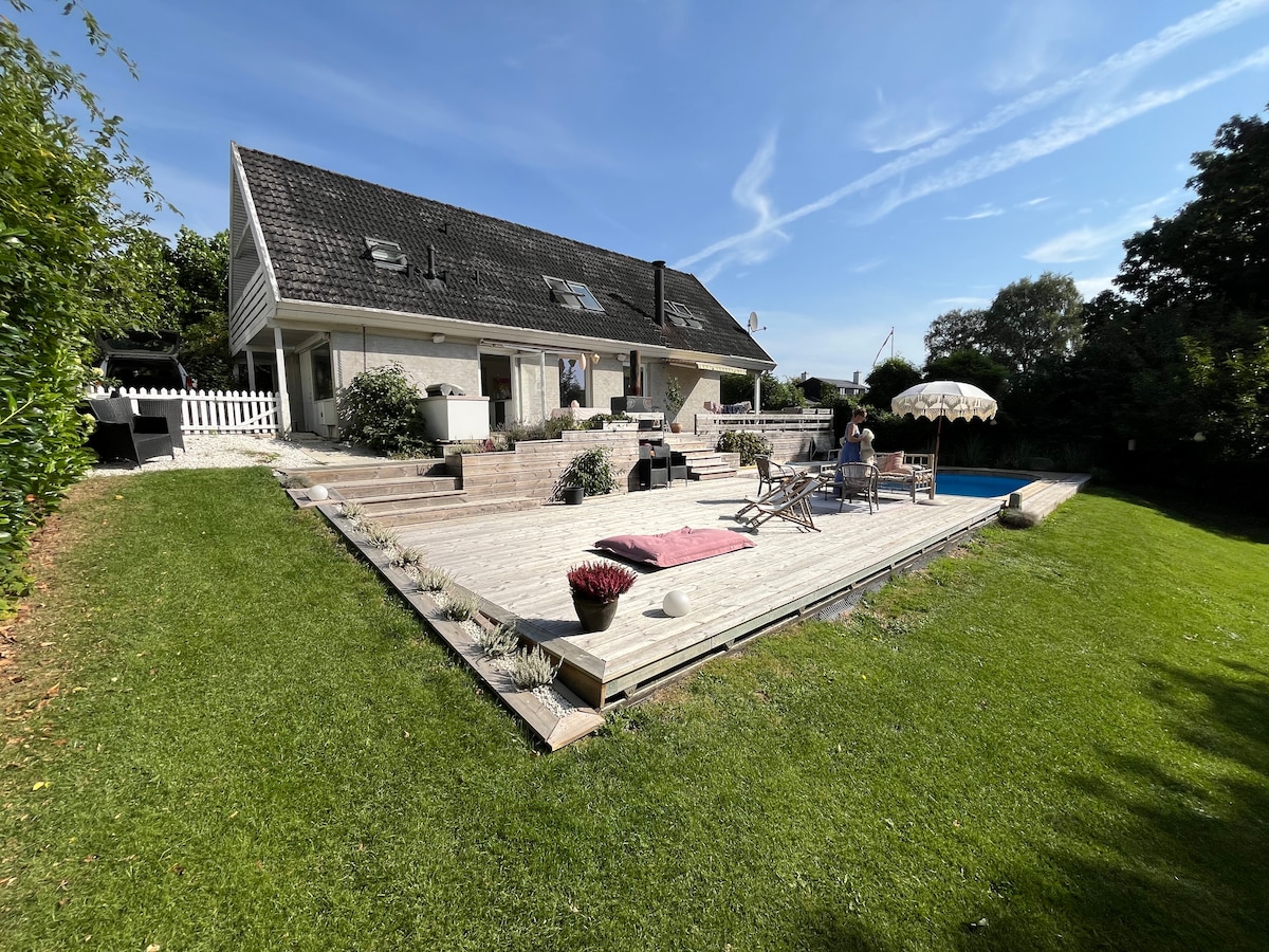 A spacious backyard area features a wooden deck with sun loungers and a patio umbrella. The house, with its sloped roof, offers large glass doors leading to the outdoor space, surrounded by green grass and well-kept shrubs.